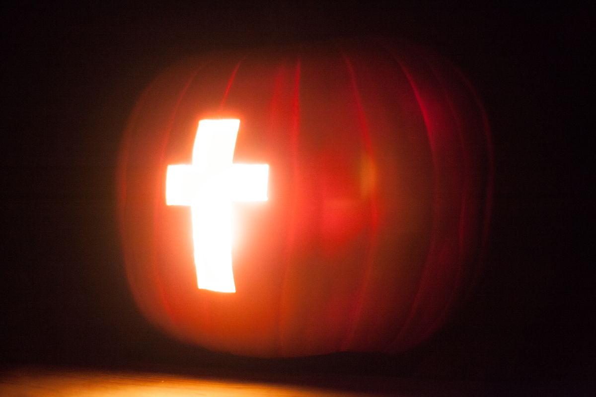 A pumpkin carved with a cross shape, illuminated from within, set against a dark background.