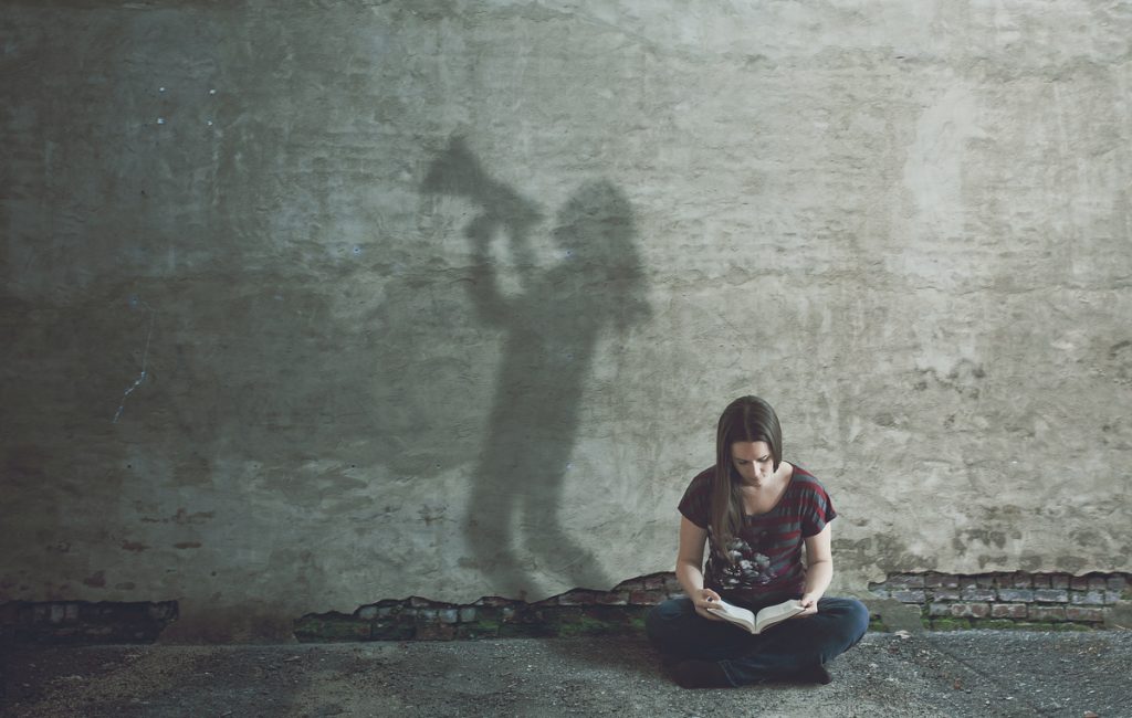 A woman sitting on the ground reading a book, with a shadow of a figure holding a megaphone projected on the wall behind her.