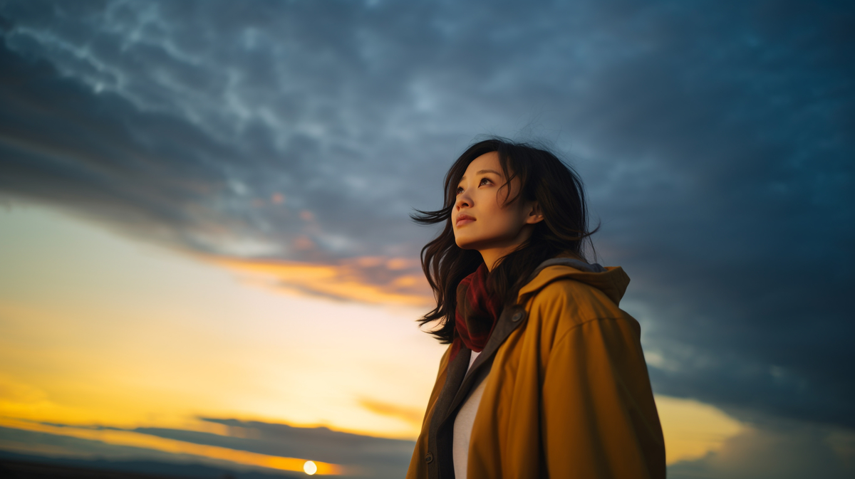 A thoughtful woman gazing upward at a colorful sunset, wearing a yellow jacket and a dark scarf against a dramatic sky.