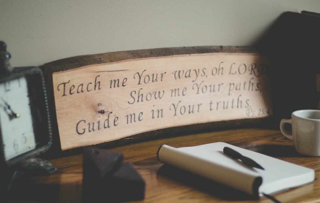 A wooden sign with the text 'Teach me Your ways, oh LORD. Show me Your paths, Guide me in Your truths. Ps. 25:4.' on a desk with a clock, a notebook, a pen, and a cup.